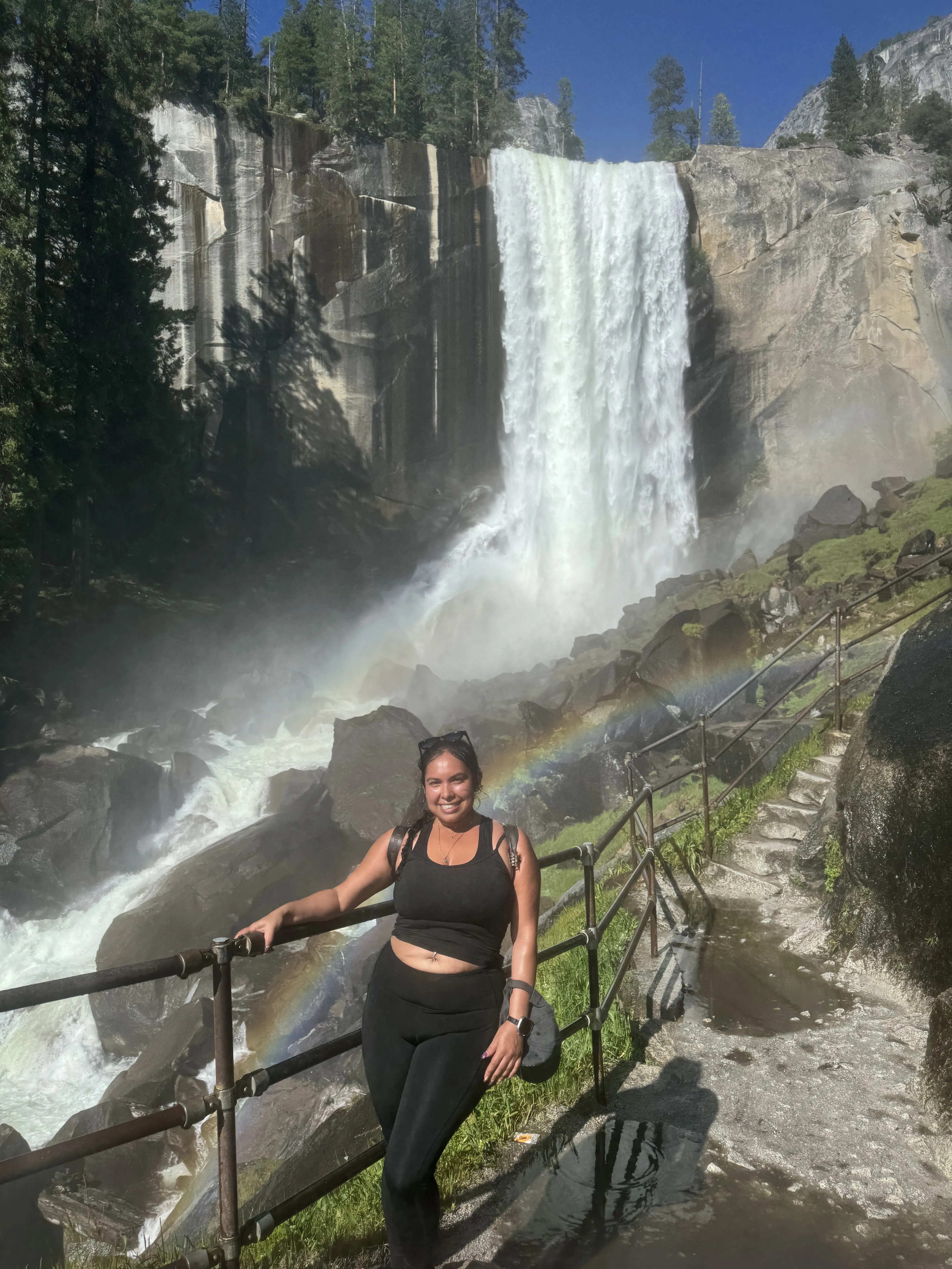 Woman standing by a waterfall with a rainbow, surrounded by trees and rocks.