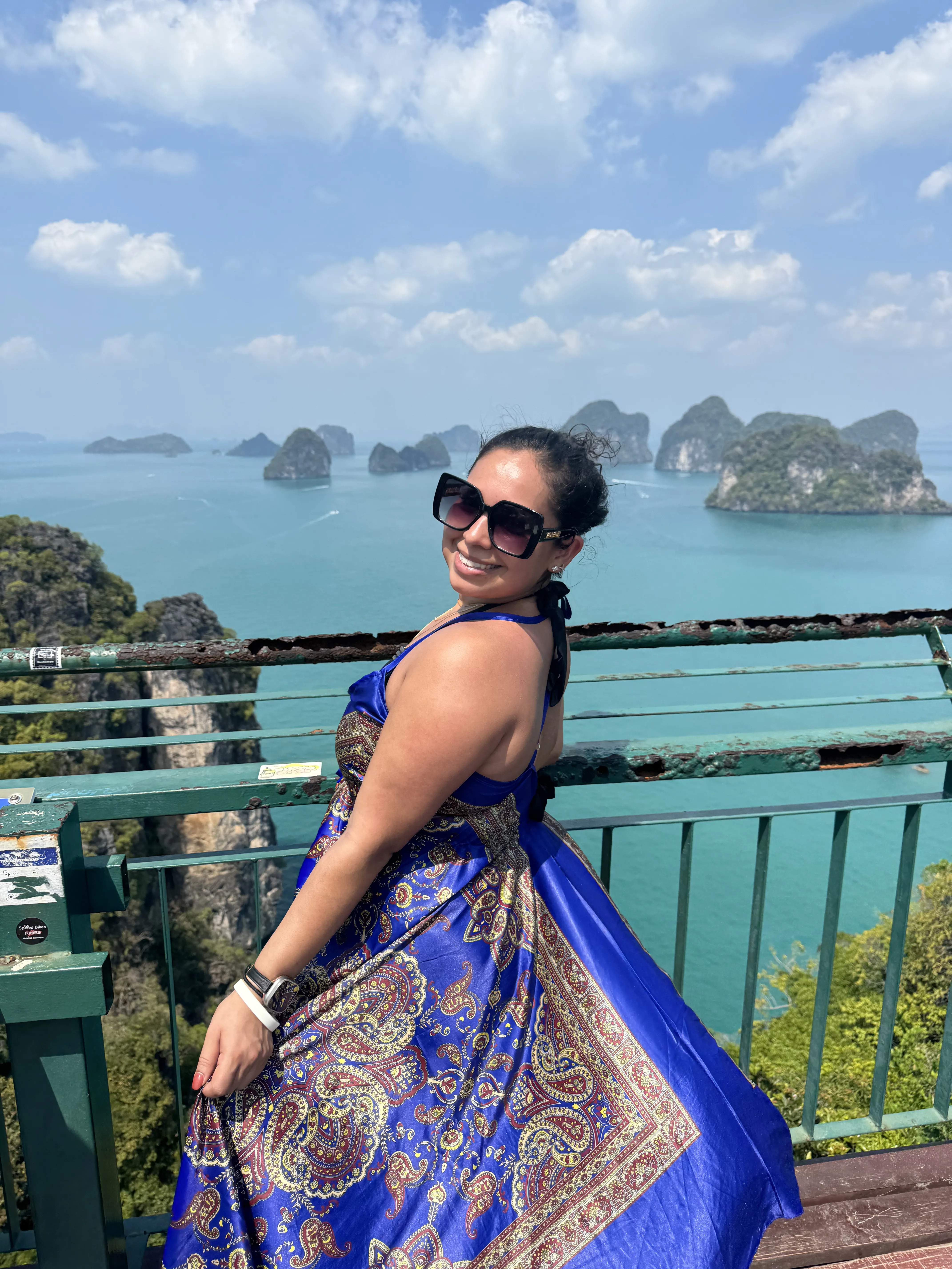 Woman in a blue patterned dress poses against a scenic coastal backdrop with islands.